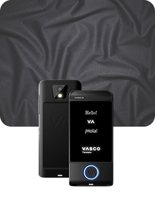 Une image de présentation du produit montrant un appareil Vasco Translator noir sous deux angles différents, sur un fond en tissu sombre et texturé. L'appareil de gauche présente une vue arrière, mettant en évidence l'objectif de l'appareil photo avec son flash et le logo « VA » en relief. L'appareil de droite présente une vue de face avec son écran allumé, affichant les messages « Hello ! », « VA », « ¡Hola ! » et le logo « VASCO Translator ». Un bouton circulaire situé au bas de la face avant de l'appareil est souligné d'une lumière bleue brillante. L'arrière-plan est constitué d'un tissu drapé gris anthracite présentant une texture subtile semblable à un maillage.