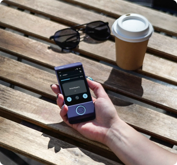 La main d'une personne tenant un petit appareil de traduction portable violet au-dessus d'une table à lattes en bois. L'écran affiche une leçon de langue avec le mot « Amerikaans ». Une tasse de café et des lunettes de soleil sont visibles à l'arrière-plan.