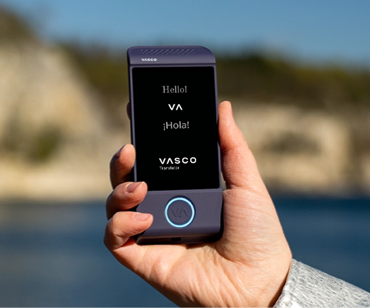 Gros plan sur une main tenant le Vasco Translator M4, un appareil de traduction électronique de couleur violette, sur fond extérieur lumineux. L'écran affiche les formules de salutation « Hello ! », « ¡Hola ! » et le logo « VASCO Translator ».